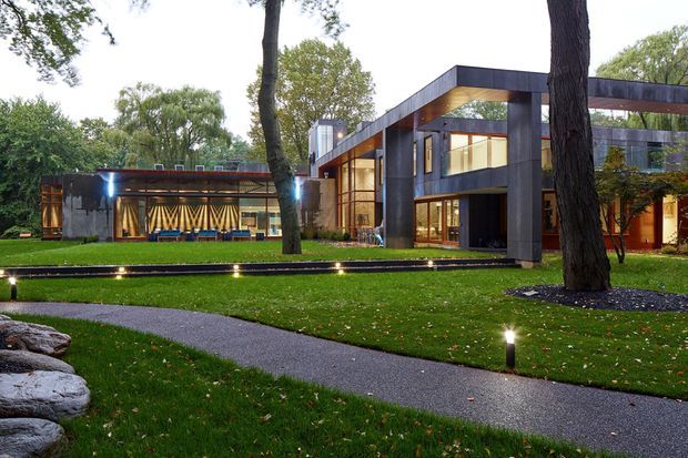 Exterior view of a large contemporary modernist residence set within a landscaped garden. The building features dark rectilinear structural frames, extensive timber-framed glazing, warm timber soffits and interior finishes, and a low horizontal wing with patterned screening. A paved garden path, lawn, mature trees, landscape lighting and stone edging occupy the foreground.