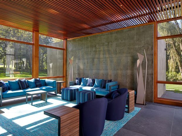 A bright, spacious modern sunroom or enclosed outdoor lounge with floor-to-ceiling glazing, exposed concrete walls, warm timber window frames, and a slatted timber ceiling. The room contains blue sofas and armchairs arranged around low timber tables, striped ottomans, and a blue area rug. Strong sunlight creates patterned shadows across the floor, with landscaped grounds visible through the windows.