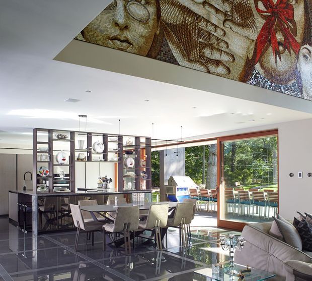 Open-plan contemporary dining and living area with a large dining table, upholstered chairs, extensive built-in open shelving, glossy dark tiled flooring, and full-height timber-framed glazing opening to an outdoor dining area. A striking mosaic artwork spans the ceiling above the room, while the bright white ceiling and abundant glazing create a spacious, polished interior.
