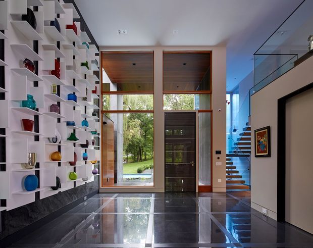 A dramatic modern residential entry hall with a highly polished dark stone floor, a large sculptural white display wall filled with colourful objects, and tall timber-framed glazing beside a timber-clad entry door. A floating open-riser stairway with a glass balustrade is visible to the right, and a framed artwork hangs on a pale wall. The double-height-feeling entrance is bright, refined, and strongly connected to the landscaped outdoors.