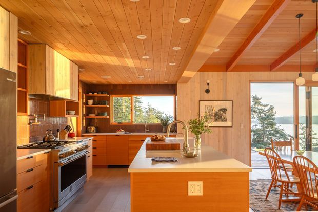 Warm, wood-lined open-plan kitchen and dining area with extensive timber cabinetry, a central island with a pale benchtop, stainless-steel cooking appliances, open shelving, and a timber dining table beside large windows. Exposed timber ceiling beams and wall panelling create a mid-century-inspired interior, with views of trees and water beyond the glazing.
