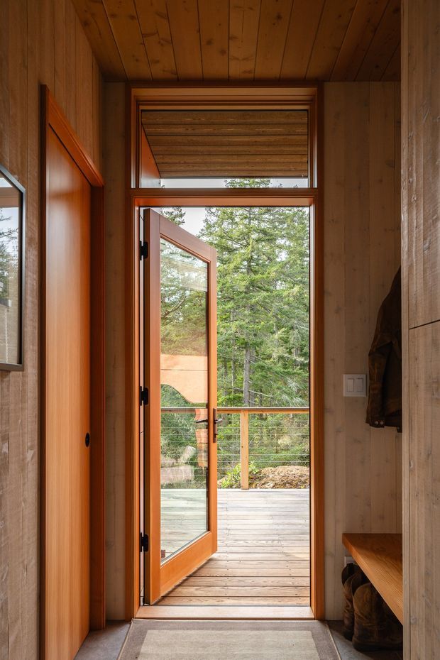 A narrow timber-lined entryway opens through a glazed wood door onto a raised outdoor deck surrounded by trees. Warm wood panelling covers the walls and ceiling, with built-in timber storage and bench seating along the sides. A small horizontal window above the door and a grey floor mat complete the welcoming cabin-like entrance.