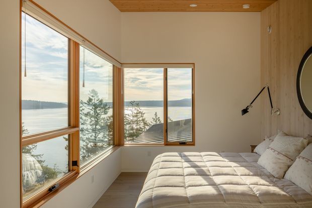 A warm, compact bedroom with a large corner arrangement of timber-framed windows overlooking a calm waterfront landscape. The room features a low-profile bed with a light quilted duvet and neutral pillows, pale walls, timber ceiling boards, timber wall panelling, and simple black wall-mounted reading lights. Natural light and expansive glazing create a serene, airy atmosphere.