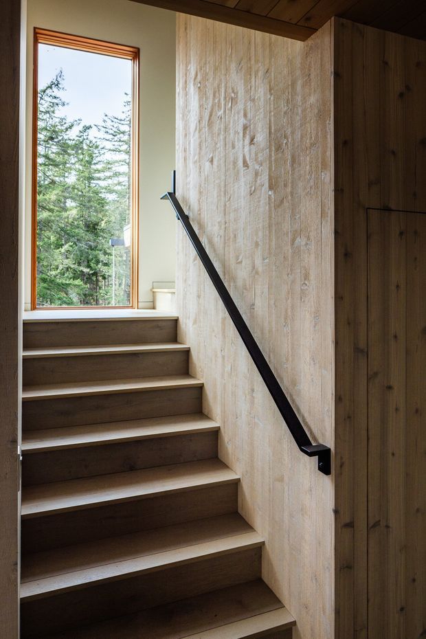 Interior view of a compact timber staircase ascending beside a large vertical window. The stair treads, risers, ceiling, wall panelling and built-in joinery are predominantly wood, with a slim matte-black handrail contrasting against the pale timber wall. The window frames a wooded outdoor setting and brings natural light into the stairwell.
