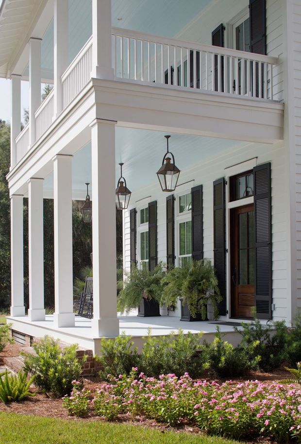Exterior view of a large white-clad house with a two-level covered front porch. The porch features tall square columns, white balustrades, pale blue beadboard-style ceilings, hanging lantern lights, dark louvered shutters, and glazed timber doors. Brick foundation details and landscaped garden beds with flowering plants occupy the foreground.