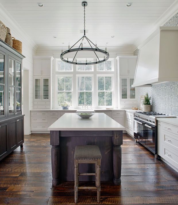 Bright, spacious traditional kitchen with a large central island, white perimeter cabinetry, dark timber island base, wide-plank timber flooring, white beadboard ceiling, extensive multi-pane windows, and a textured light mosaic tile backsplash. A large black metal ring pendant hangs above the island, while a freestanding stainless-steel range and hood occupy the right side. Glass-front cabinetry and woven baskets add visual detail.