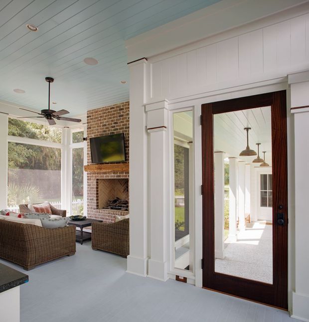 Bright enclosed sunroom or living area with white-painted vertical wall panelling, a pale blue-white beadboard ceiling and matching painted floor. The room features expansive glazing, wicker seating, a brick fireplace with television above it, and a dark timber glazed entry door opening toward a columned exterior porch or walkway. The traditional detailing, abundant daylight and connection to outdoors create a relaxed coastal character.