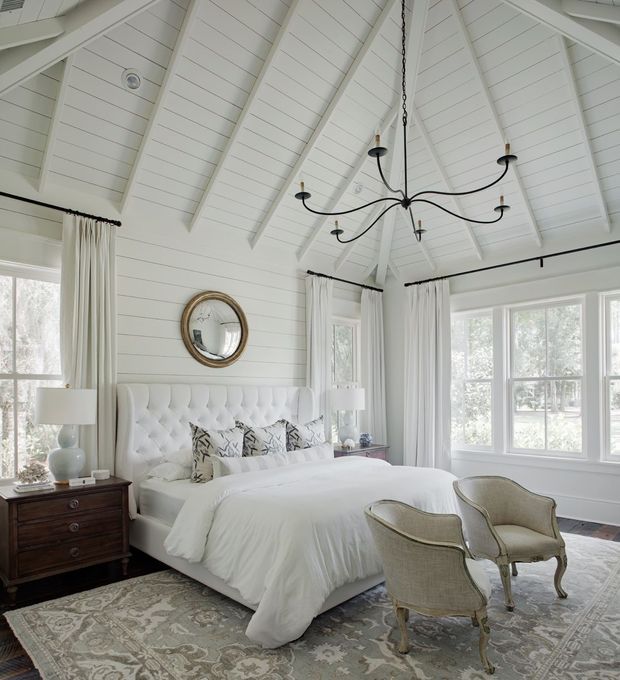 Bright, elegant bedroom with a white upholstered tufted bed, white bedding, bedside tables and lamps, patterned area rug, upholstered occasional chairs, and tall curtained windows. The room features white horizontal wall panelling and a dramatic vaulted ceiling with exposed white rafters and a black multi-arm chandelier.