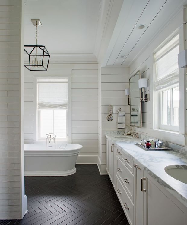 Bright, refined bathroom with white horizontal wall panelling, a freestanding bathtub beneath a window, and a long white vanity with marble-patterned benchtop, integrated basin, and polished metal tapware. A large mirror, wall sconces, folded towels, and a black geometric pendant complement the dark herringbone floor. The room has abundant natural light and a classic coastal feel.