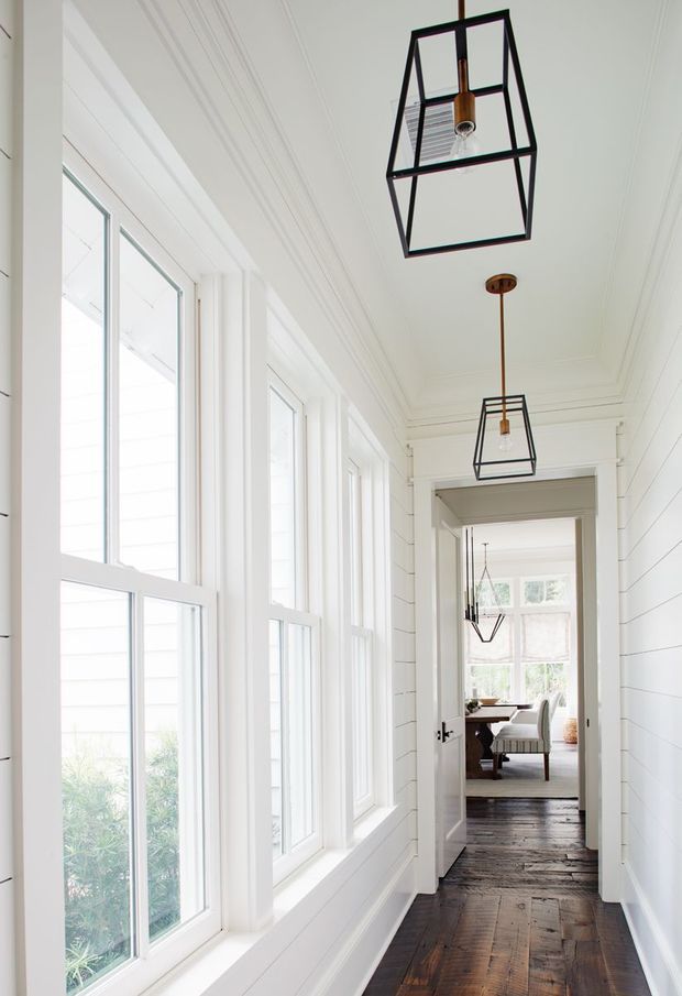 A bright, narrow residential hallway with full-height white-framed windows along one side, white horizontal wall panelling, ornate ceiling mouldings, dark reclaimed timber flooring, and a series of black geometric pendant lights. The hallway opens through a doorway to a dining area visible in the distance.