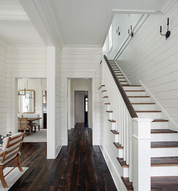 Bright, narrow residential hallway and stairway with white horizontal wall panelling, crisp decorative ceiling trim, dark distressed timber floors, and a staircase with white balusters, dark timber handrail, and dark wood treads. A dining area is visible through the open doorway on the left, while a corridor extends beneath the stairs. Black wall-mounted candle sconces line the stair wall, creating a refined coastal-traditional interior.