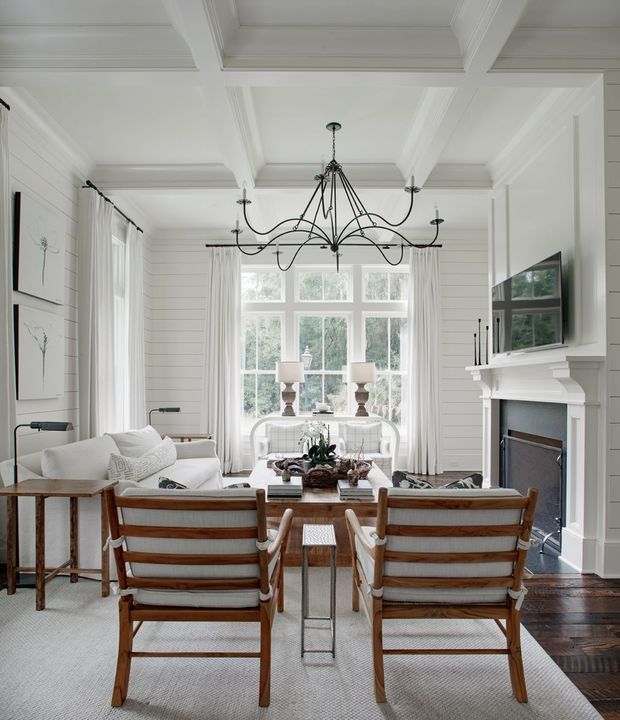 Bright, refined open-plan living and dining room with white shiplap walls, a coffered ceiling, tall multi-pane windows, and dark timber flooring. A large black metal chandelier hangs over a dining table surrounded by upholstered timber-framed chairs. The room also includes a white sofa, pale area rug, fireplace with a substantial white mantel, wall-mounted television, and light neutral furnishings.