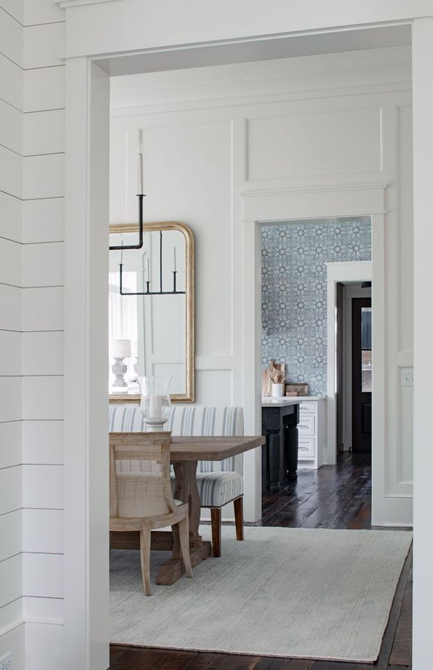 A refined transitional interior view through a wide, heavily trimmed opening. The foreground shows a dining area with a rustic wood table, woven dining chairs, and a striped upholstered bench beneath a large rounded-top mirror and simple black linear pendant. Beyond it is a dark timber-floored space with patterned blue-grey wallpaper, white cabinetry, and classic wall panelling. The palette is predominantly white with natural timber, soft blue-grey, and black accents.