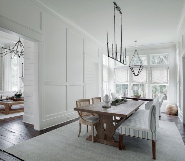Bright, refined dining room with white panelled walls, a large rustic timber dining table, upholstered dining chairs and bench seating, and a pair of striking black linear and geometric pendant lights. Tall bay-style windows with Roman blinds provide abundant natural light, while dark timber flooring and a large pale rug add contrast. A living area is visible through the wide opening to the left.