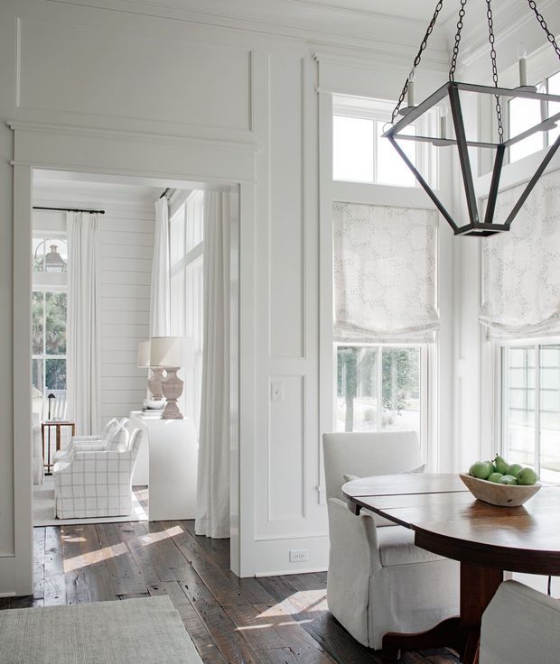 Bright, refined dining area in a traditional home, featuring a dark timber dining table with upholstered slipcovered chairs, tall multi-pane windows with patterned Roman shades, white paneled walls and trim, and a large black geometric pendant chandelier. An adjoining sitting room with white furnishings is visible through the wide opening.