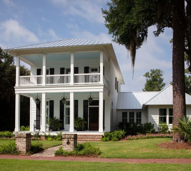 Exterior view of a large white two-storey house with a covered upper and lower front veranda, white balustrades, square columns, horizontal weatherboard cladding, dark shutters and doors, and pale metal pitched roofs. A lower side wing connects to the main volume. Brick porch piers, a brick path, manicured planting and mature trees complete the residential streetscape.