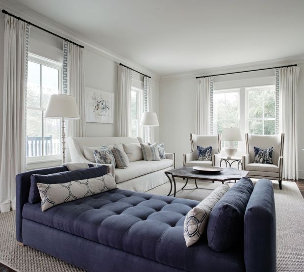 A bright, elegant living room with large windows, white walls, layered curtains, a cream sofa, two upholstered armchairs, and a deep navy tufted chaise-style sectional in the foreground. Decorative patterned cushions, a round coffee table, table lamps, and a neutral rug create a refined, comfortable interior. The room has traditional architectural detailing with contemporary furnishings and a restrained navy, white, and grey palette.
