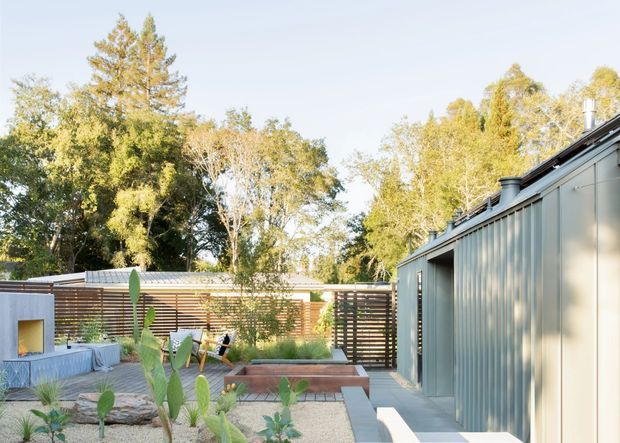 A contemporary single-storey courtyard-style exterior with a long corrugated metal-clad building on the right, a paved outdoor seating area, raised garden beds, timber privacy screens, and a small built-in outdoor fireplace or barbecue structure. Mature trees surround the property, creating a leafy suburban or bush setting. The composition is bright and relaxed, with strong indoor-outdoor living cues.