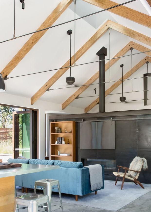 Bright open-plan living room with a vaulted white ceiling, exposed timber roof beams, suspended black pendant lighting, and a prominent dark metal fireplace surround with a black flue. A blue sectional sofa faces the fireplace, with a rocking chair, built-in timber shelving, large glazed opening, and polished concrete flooring. The room combines warm timber, cool blue upholstery, and industrial metal detailing.