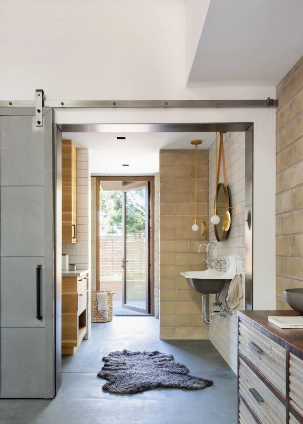 A bright industrial-style bathroom or washroom viewed through a large sliding barn-style door. The space features exposed tan masonry block walls, white rectangular wall tiles, a polished concrete floor, a wall-mounted pedestal sink with exposed chrome plumbing, a round mirror, and hanging globe pendant lights. Timber cabinetry and a timber-fronted vanity are visible along the sides, with a glazed exterior door at the far end.