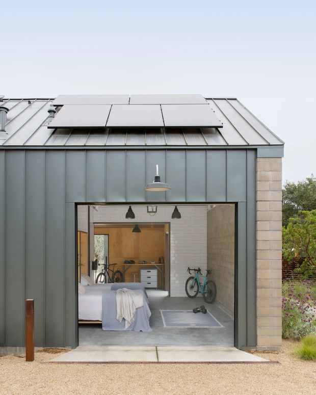 Exterior view of a compact modern outbuilding or converted garage with a standing-seam metal pitched roof and broad roof overhang. Dark grey vertical metal cladding frames a large open glazed or garage-style opening, revealing a combined bedroom and home gym interior with a bed, bicycle, exercise mat, and built-in timber cabinetry. Pale masonry side walls and a restrained industrial-modern palette are visible, set beside landscaped planting and gravel paving.
