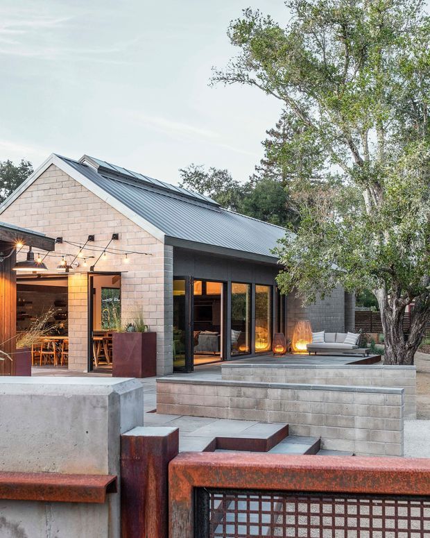 Exterior view of a contemporary single-storey house with a steep gabled metal roof, pale masonry block walls, extensive dark-framed glazing, and a partially enclosed outdoor entertaining area. Broad stepped masonry terraces, concrete and weathered steel site elements, outdoor seating, decorative lighting, and mature trees define the approach and courtyard-like outdoor space.