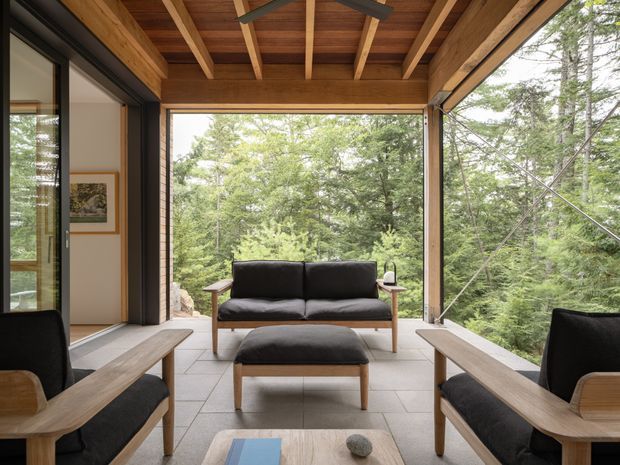 A covered, open-sided living lounge surrounded by forest, with black upholstered mid-century-style sofas and chairs, timber armrests and legs, a matching ottoman, and a low timber coffee table. Large glazed openings connect the space to the wooded landscape. The ceiling has exposed timber beams and warm wood lining, while the floor is finished in large light-grey tiles. The space feels calm, natural, and strongly connected to the outdoors.