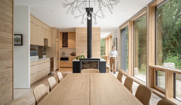 A bright open-plan kitchen and dining space with extensive light timber cabinetry and wall panelling, a long timber dining table, and a central black fireplace or stove with a flue. Full-height timber-framed glazing runs along the right side, opening views to a wooded outdoor setting. A sculptural branch-like pendant light hangs above the dining area. The room has a warm, refined contemporary character with strong indoor–outdoor connection.
