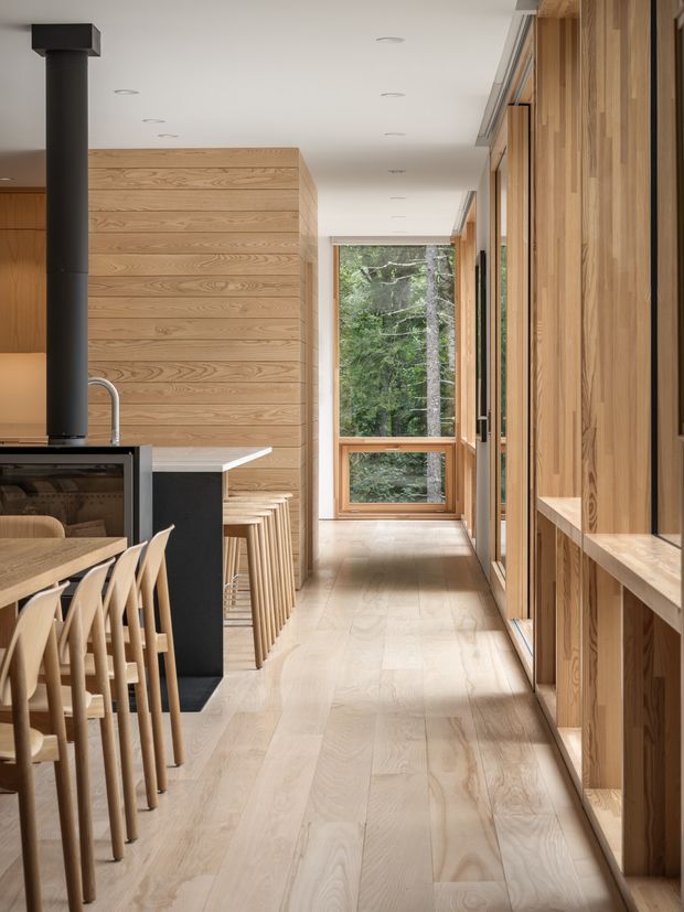 A warm, light-filled residential interior showing a long circulation space beside an open kitchen and dining area. Pale timber flooring, extensive vertical timber wall panelling and built-in joinery create a cohesive natural material palette. A dark kitchen island and black wood-burning stove provide contrast, while a row of pale timber dining chairs lines the foreground. Full-height timber-framed glazing at the end of the corridor overlooks dense forest.