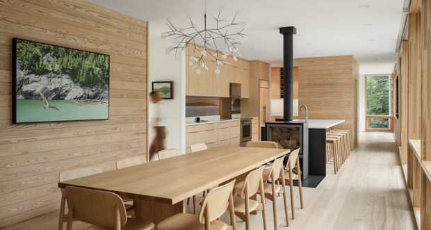 Open-plan kitchen and dining area with extensive light timber wall panelling and cabinetry. A long timber dining table with matching chairs occupies the foreground, while a kitchen island with bar seating extends toward the rear. A black freestanding wood-burning stove provides a strong contrast, complemented by a sculptural branch-like pendant light and large windows overlooking greenery.