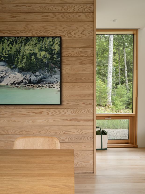 Warm contemporary interior featuring a full-height natural wood wall with prominent horizontal grain, a framed forest landscape artwork, and a simple light-wood dining or work table with matching chair. A timber-framed vertical window reveals a wooded setting, while pale timber flooring and neutral plaster walls create a calm, nature-connected atmosphere.