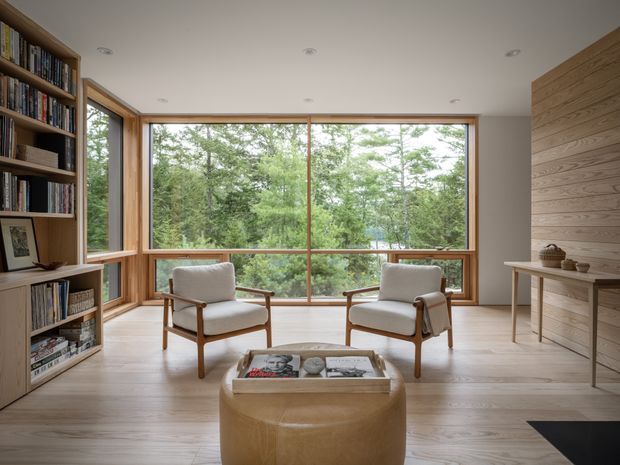 A calm, light-filled living room with extensive timber-framed glazing overlooking a wooded landscape. Built-in timber bookshelves line the left wall, while a horizontal timber feature wall and slim console occupy the right. Two upholstered lounge chairs face a rounded timber coffee table on pale timber flooring, creating a warm, minimalist retreat.