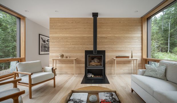 A bright, refined living room with extensive timber-framed corner glazing overlooking trees. The room features a horizontal light-wood feature wall, a central black freestanding wood-burning fireplace with flue, pale timber flooring, a light sofa, mid-century-style armchairs, and simple console furniture. The space has a calm natural connection to the surrounding landscape and a standard-height ceiling.