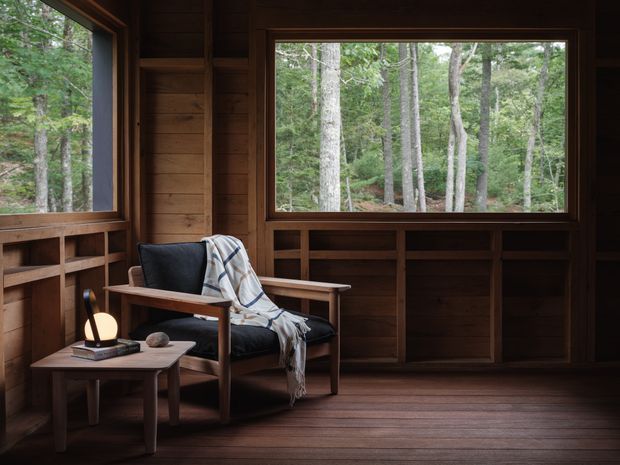 A serene timber-lined reading nook with a dark upholstered lounge chair, draped throw, small wooden side table and lamp, and extensive windows looking into a wooded landscape. Built-in timber shelving and cabinetry run beneath and beside the windows, while dark timber floorboards and wall panelling create a warm cabin-like interior.
