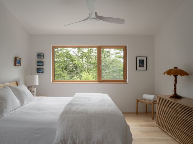 A calm, light-filled bedroom with a neatly made white bed, pale neutral walls, timber-framed picture window overlooking leafy trees, light timber flooring, a ceiling fan, and a slatted timber sideboard topped with a vintage-style lamp. Minimal artwork and restrained furnishings create a clean, understated interior.
