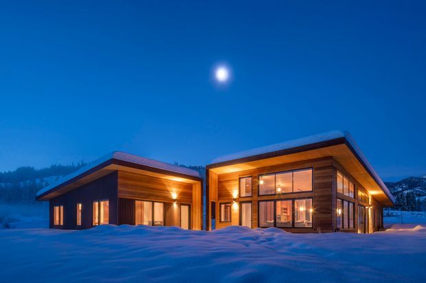 Twilight exterior view of a modern timber-clad mountain home composed of two low, gabled volumes connected by a glazed central entry. Warm interior lighting shines through large windows and glazed doors, contrasting with the deep blue sky and surrounding snow-covered landscape. The building has broad roof overhangs, dark wood cladding, and a minimalist alpine lodge character.