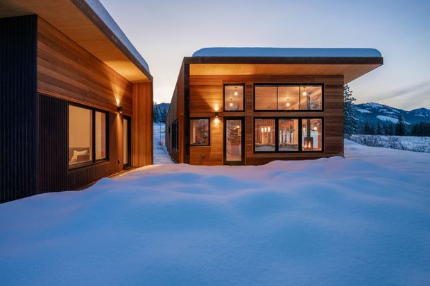 Exterior view of a compact modern timber-clad building complex set in deep snow, with two low-profile pavilion-like volumes connected by a narrow passage. The buildings feature warm wood siding, dark vertical cladding accents, extensive black-framed glazing, and gently curved or projecting flat roof forms. Interior lighting glows through the windows at dusk, with a forested mountain landscape visible in the background.