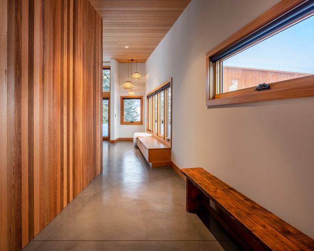 A long, narrow modern hallway with a polished concrete floor, warm vertical timber wall panelling, a matching timber ceiling, and timber-framed windows. Built-in timber benches and cabinetry line the corridor, while simple pendant lights create a warm, refined atmosphere.