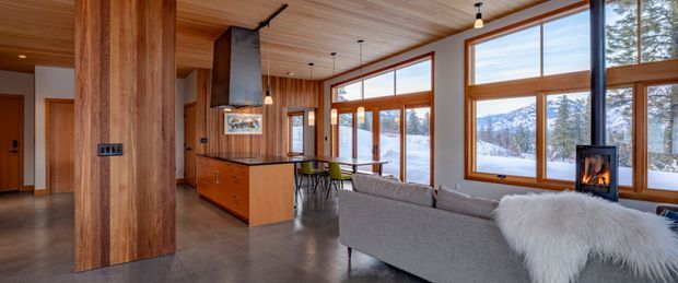 Open-plan contemporary living and dining area with a warm timber-lined ceiling and substantial timber structural elements. A wood-burning fireplace, kitchen island, dining table, and large timber-framed windows overlook a snowy forested mountain landscape. The room has polished grey concrete flooring, restrained furnishings, and abundant natural light.