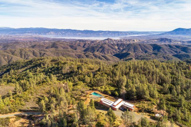Aerial view of a secluded low-rise building complex nestled among dense trees and rugged, rolling mountain terrain. The surrounding landscape dominates the image, with a visible turquoise swimming pool and several connected pale-roofed structures near a winding access road. Bright daylight and expansive layered ridgelines create a remote, scenic setting.