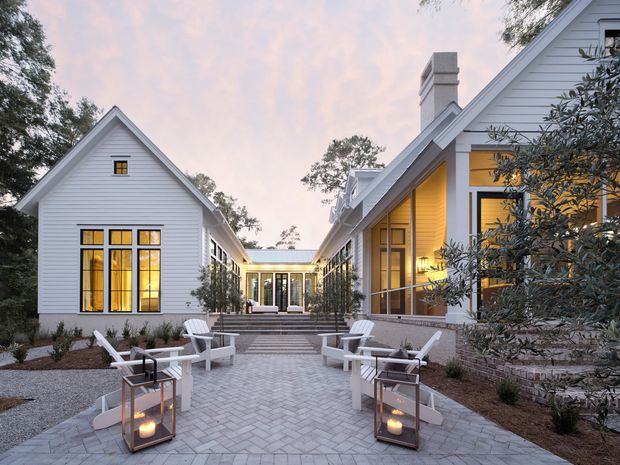 A landscaped courtyard between several connected white weatherboard buildings with steep gabled roofs. Tall black-framed windows glow warmly from within, while a pale herringbone-paver path leads through the courtyard past white outdoor lounge chairs and portable lanterns. The composition highlights the residential or hospitality-style building exterior, covered link, planting, and inviting evening atmosphere.
