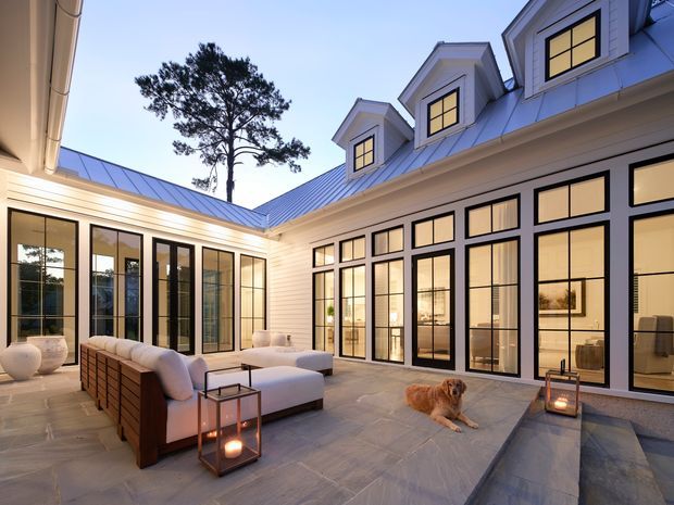 A refined courtyard terrace enclosed by a substantial light-coloured home with a blue-grey metal roof, repeated dormer windows, and extensive black-framed glazing. The paved outdoor living area contains a large upholstered sectional, lanterns, and a small fire feature, with warm interior lighting visible through the windows. A dog rests on the terrace. The composition emphasizes the landscaped courtyard, indoor-outdoor connection, and symmetrical gabled roof forms.
