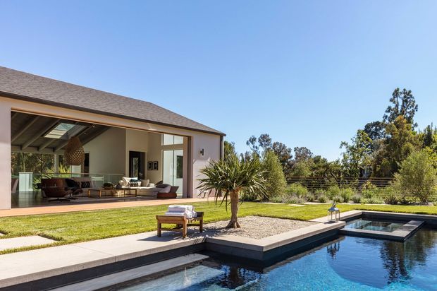Contemporary single-storey residence viewed from a landscaped backyard, with a broad low-pitched roof, expansive glazed opening to an indoor-outdoor living area, paved terrace, lawn, sculptural planting and a geometric swimming pool with raised square-edged sections.
