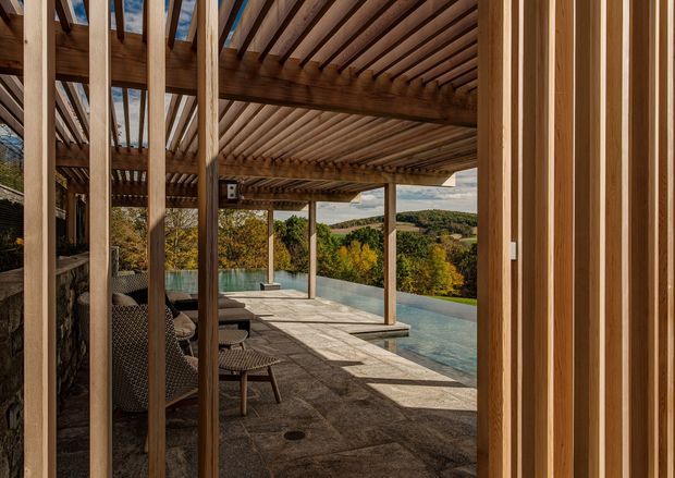 View from a timber-framed opening onto a covered outdoor terrace with a slatted timber pergola, stone paving, outdoor seating, and a narrow reflecting or swimming pool overlooking a wooded, rolling landscape. The image emphasizes warm natural timber structure and the connection between the sheltered terrace and the surrounding scenery.