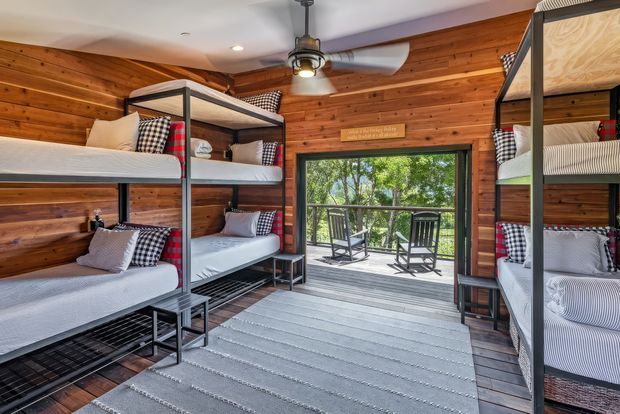 A rustic bunk room lined with warm horizontal timber boards, featuring three sets of dark metal bunk beds dressed in coordinated grey, white, red and black-check bedding. A wide glass door opens onto a timber deck with outdoor seating and leafy surroundings. The room includes a ceiling fan with integrated light, a grey striped area rug, and a compact cabin-like layout.