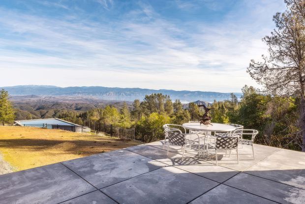 A wide outdoor terrace with large grey paving slabs and a white outdoor dining table and chairs, overlooking a landscaped hillside and distant mountain ranges. The scene is sunlit with mature trees and expansive views.