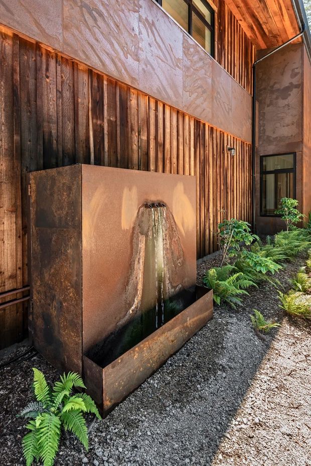 An exterior courtyard or garden passage featuring a sculptural rusted metal water feature against a dark vertical timber-clad wall. A textured rendered upper wall, black-framed windows, exposed timber soffit, gravel paving, and lush fern planting create a warm, earthy architectural setting.