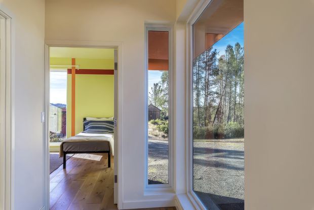 A bright residential hallway or circulation space with floor-to-ceiling corner glazing and a view to a gravelled outdoor area with trees. An open doorway reveals a bedroom with a bed, warm timber flooring, and vivid lime-green walls accented by bold red and orange trim.