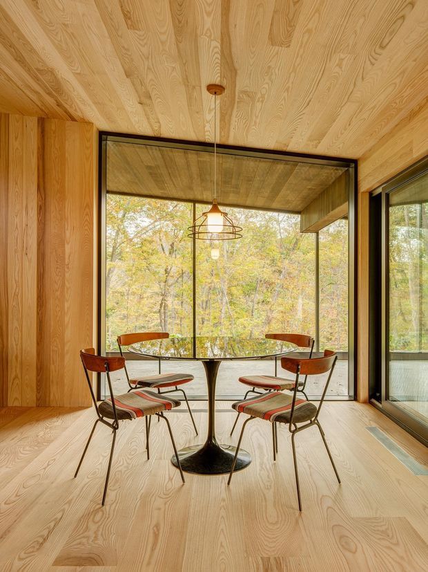 A compact timber-lined dining nook with floor, walls, and ceiling clad in warm natural wood. A round pedestal dining table is surrounded by four mid-century style upholstered chairs. Full-height black-framed glazing opens the space visually to a wooded landscape, while a delicate pendant light hangs above the table. The room has a warm, refined, nature-connected character with strong mid-century detailing.