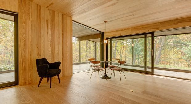 A warm, wood-lined open-plan living and dining area with extensive floor-to-ceiling glazing overlooking a wooded landscape. The room features pale timber flooring, walls and ceiling, dark-framed windows and doors, a compact mid-century-style dining set, and a dark upholstered lounge chair. Natural light and the surrounding trees create an airy, calm atmosphere.